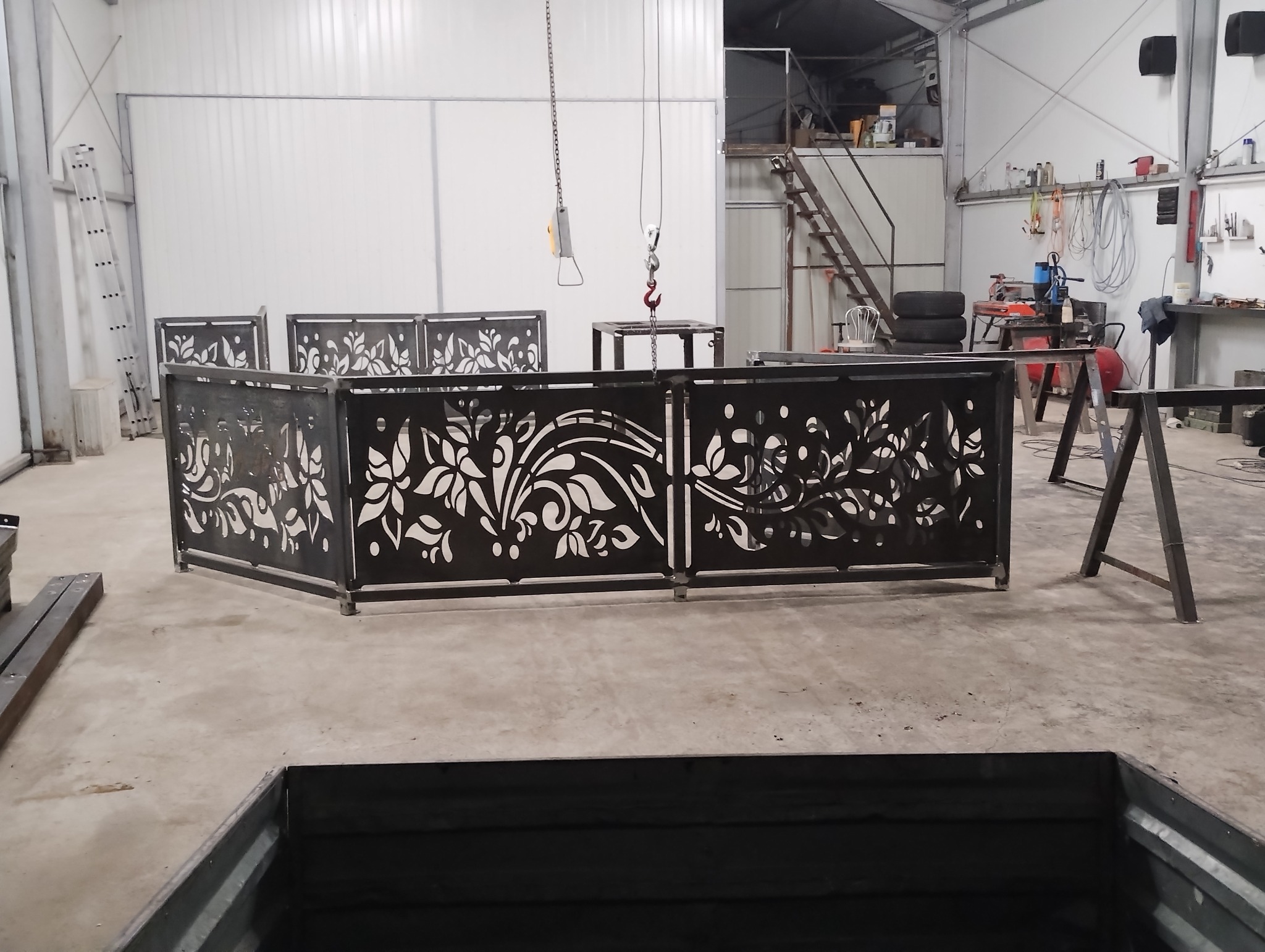 Metalowe panele dekoracyjne z wycinanym wzorem roślinnym, ustawione w hali produkcyjnej. Widoczny suwnica i narzędzia, co sugeruje etap produkcji.