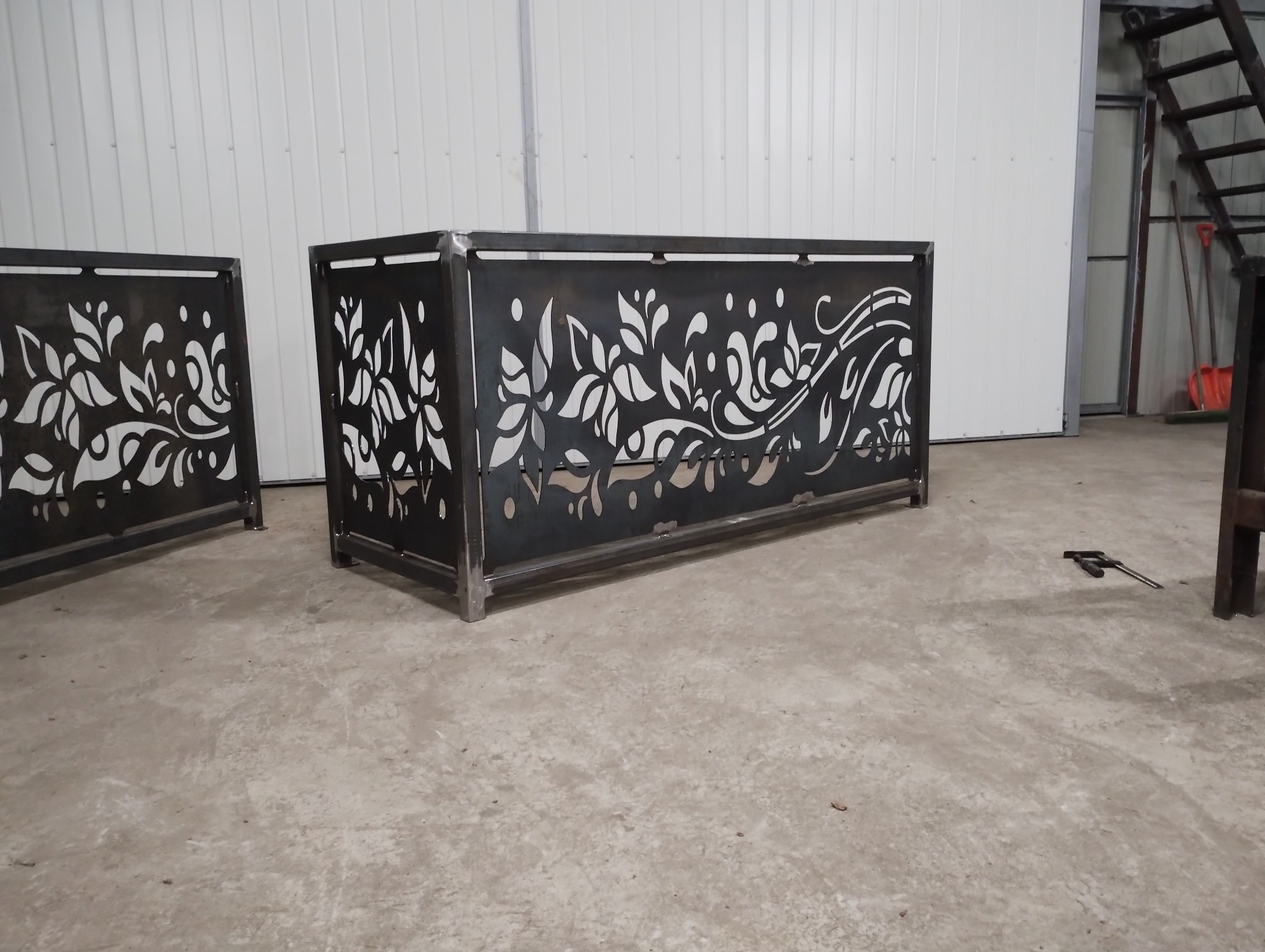 Dekoracyjny, spawany kwietnik z motywem roślinnym, wykonany z ciemnego metalu. Widoczne spawane łączenia i subtelne wykończenie. W tle biała ściana i schody.