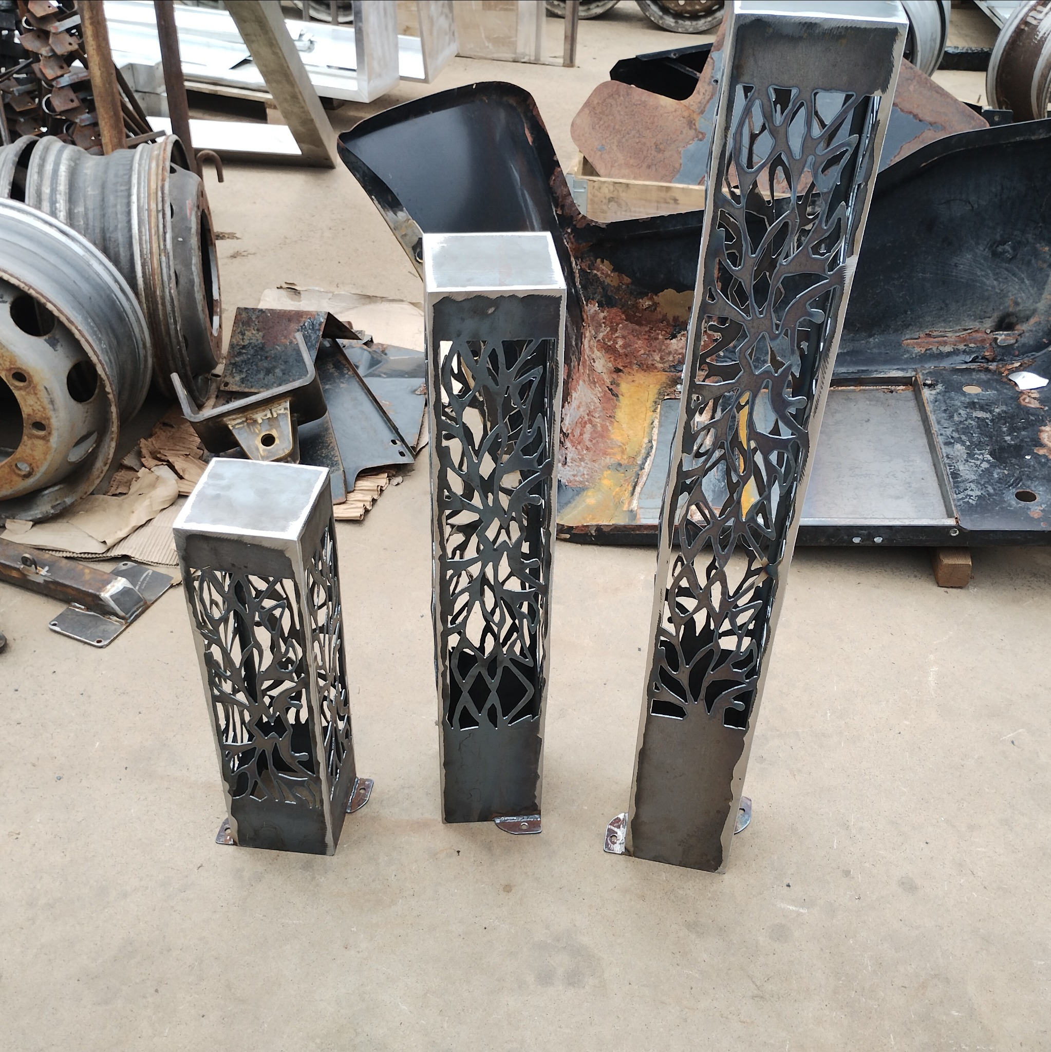 Trzy metalowe, spawane słupki z motywem drzewa, różnej wysokości, stoją na warsztatowym tle z elementami metalowymi i kołami. Prezentacja rękodzieła metalowego.