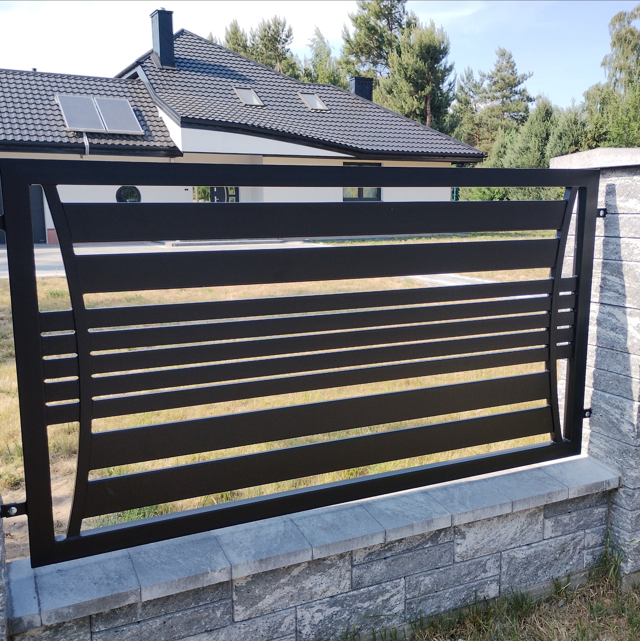 Nowoczesne, czarne, metalowe ogrodzenie panelowe z poziomymi szczeblami, zamontowane na murku z szarej kostki, na tle domu z ciemnym dachem i zieleni.