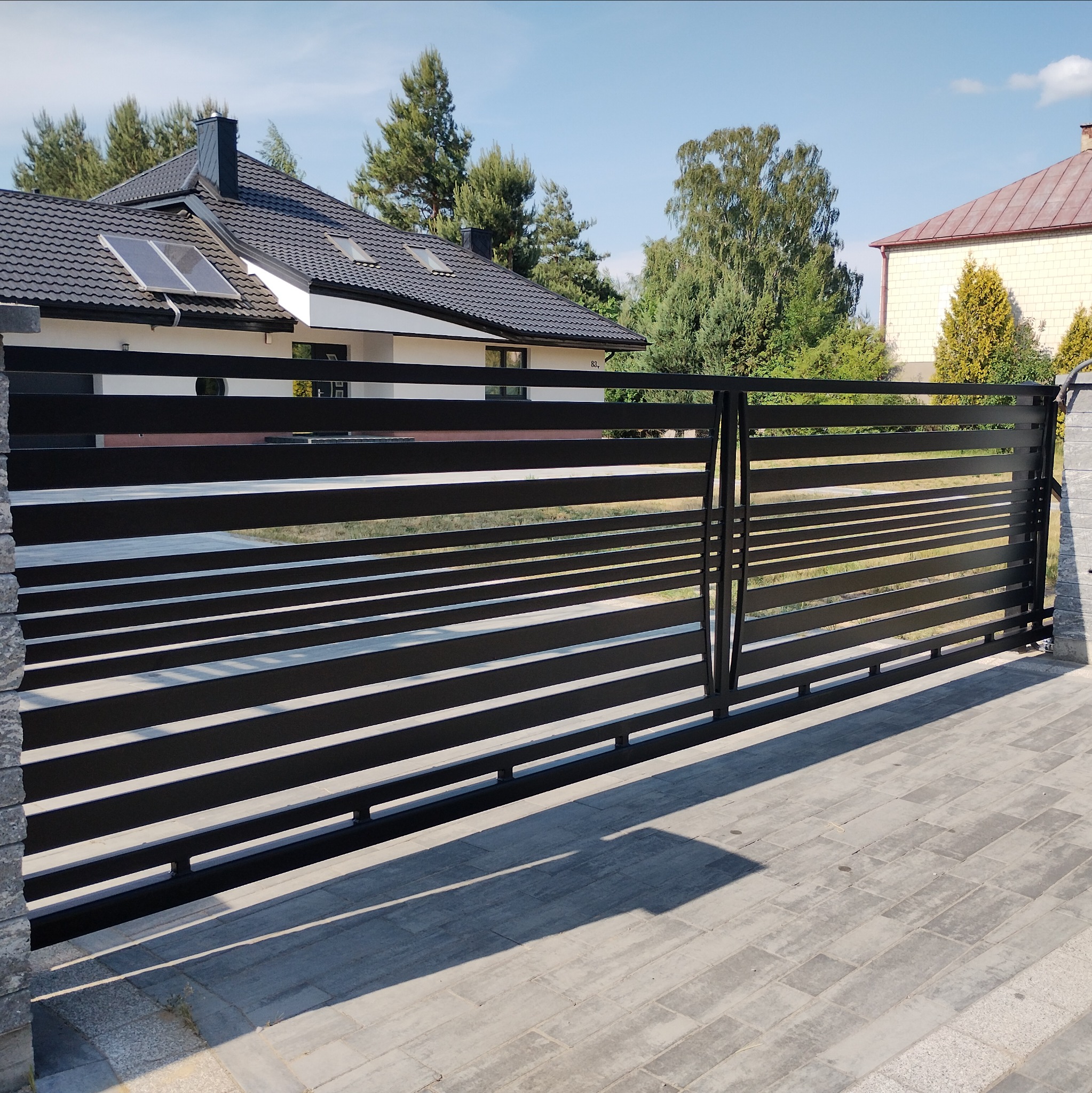 Nowoczesna, spawana brama posesyjna z poziomych profili, pomalowana na czarno, z widocznym domem w tle. Brama zamontowana na betonowych słupkach.