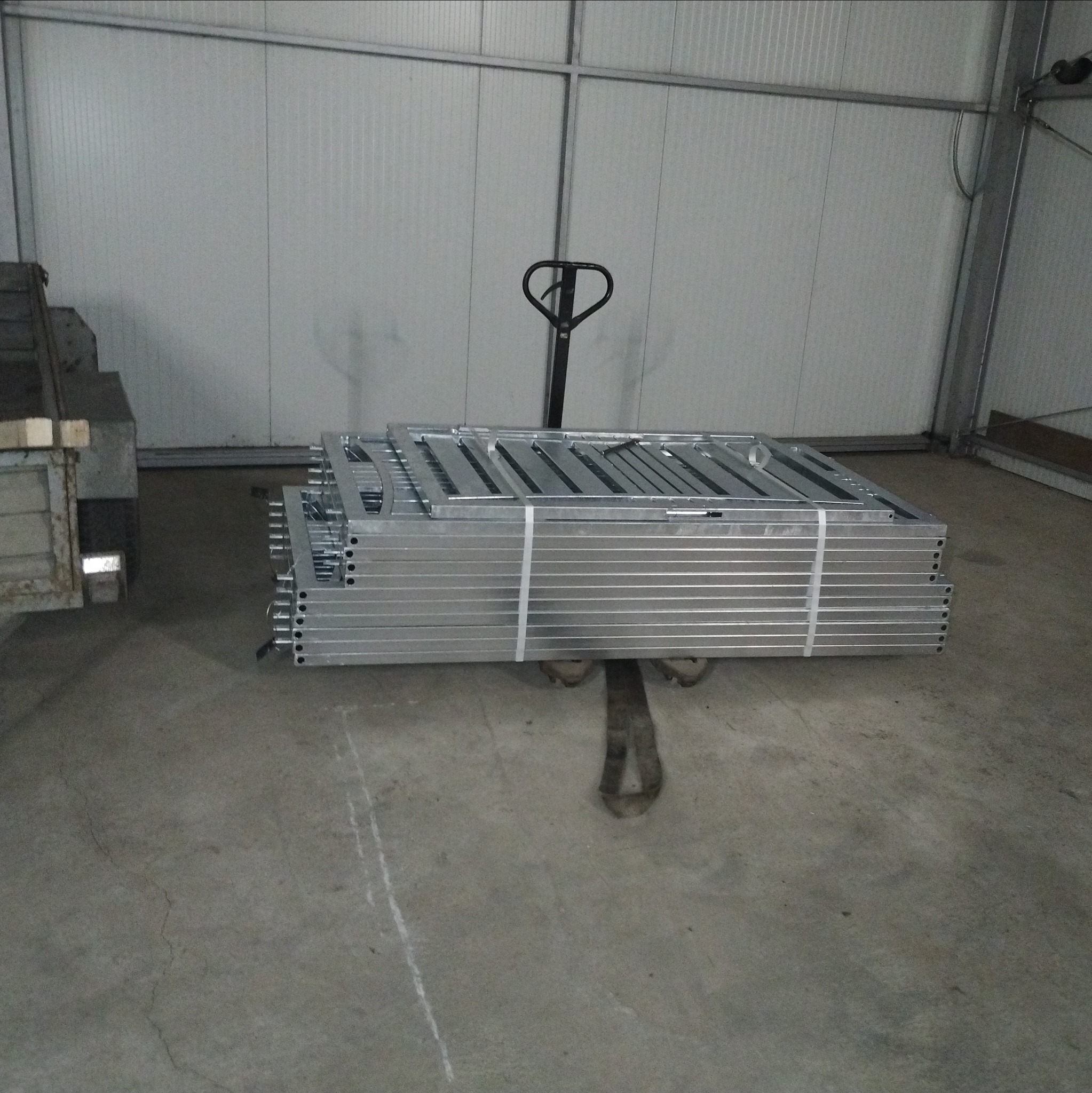 Stos srebrnych, metalowych ram spiętych taśmą, umieszczonych na wózku paletowym w hali z blachy falistej. Widoczny fragment przyczepy.