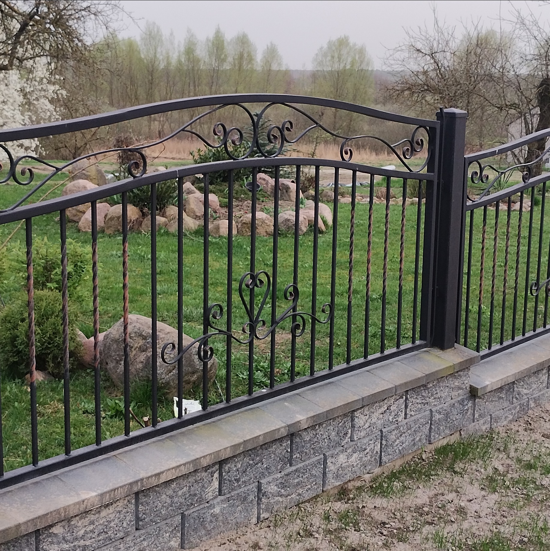 Czarny, kuty płot z ozdobnymi elementami na kamiennej podmurówce, otoczony zielenią. Elementy dekoracyjne w kształcie serca i spirali.