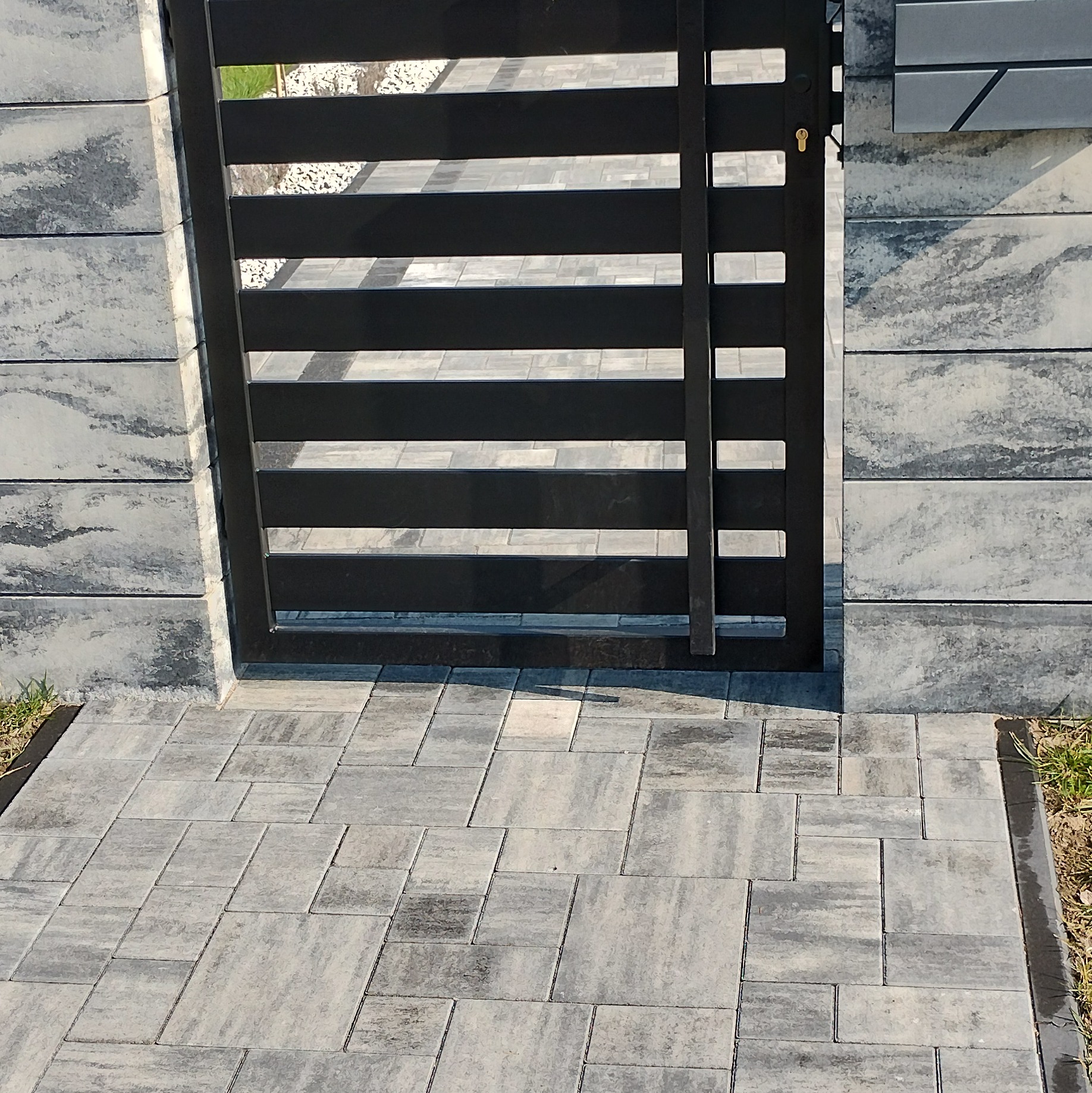 Nowoczesna, antracytowa brama z poziomych profili, zamontowana w szarym, betonowym ogrodzeniu modułowym, z dopasowaną kolorystycznie kostką brukową.