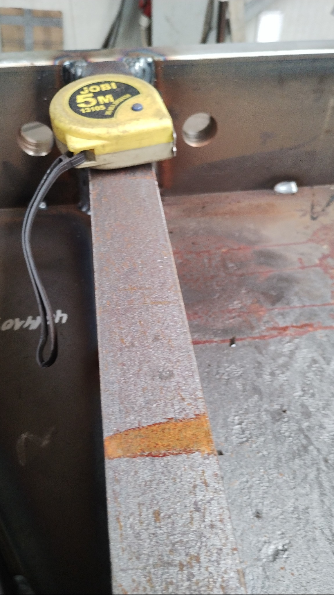 Spoina po spawaniu na metalowej konstrukcji, z żółtą miarką zwiniętą na górze. Widoczne otwory w metalu i ślady rdzy.