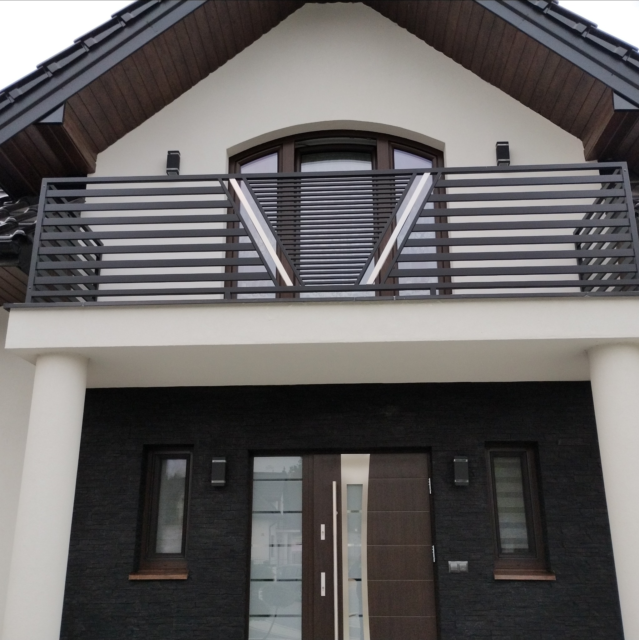 Nowoczesna, spawana balustrada balkonowa z poziomych profili, z geometrycznym wzorem w kształcie litery V, na tle elewacji domu z ciemnej cegły i białych kolumn.