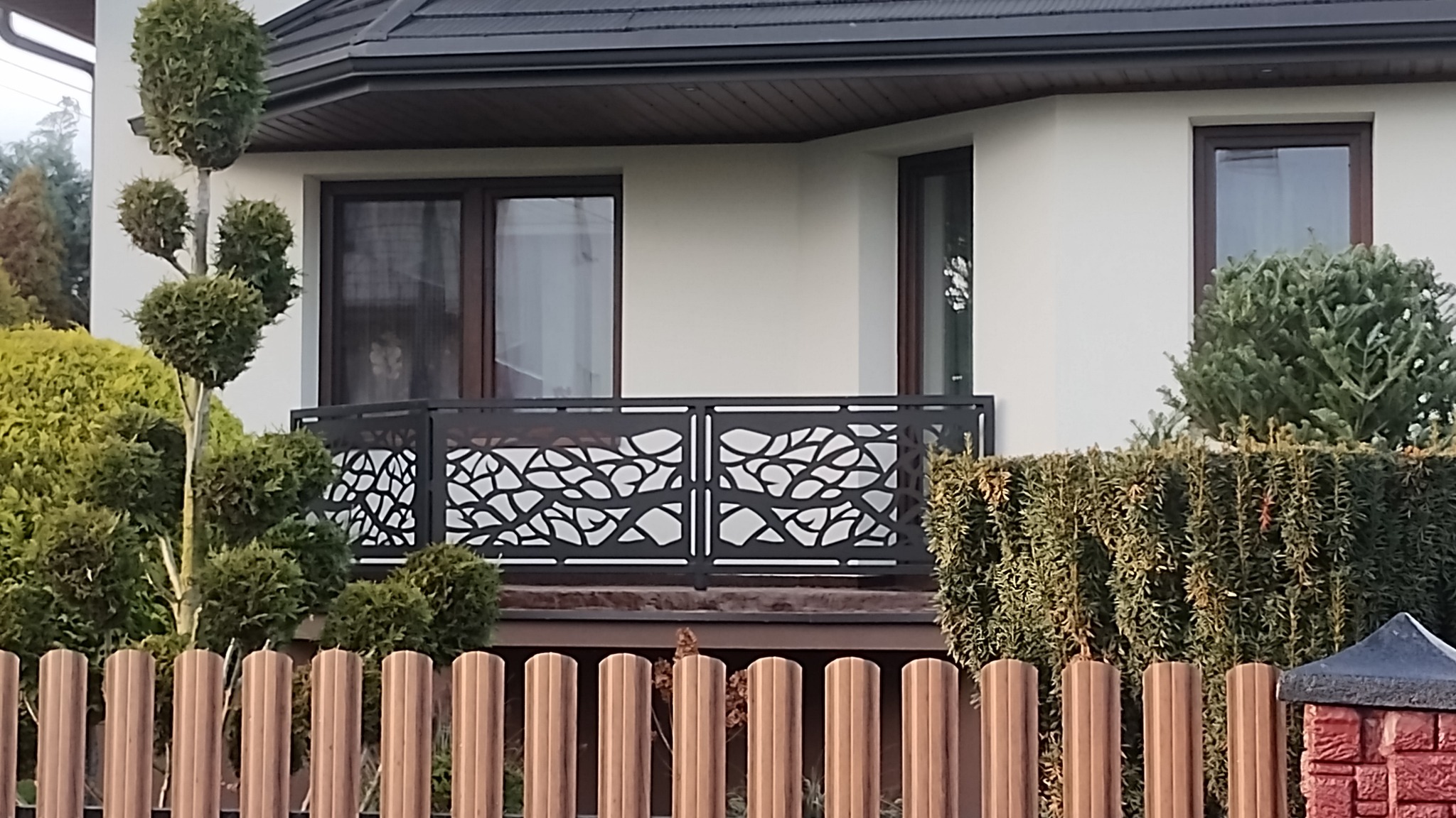 Balkon z dekoracyjną, ażurową balustradą w motywie liści, widoczny zza ogrodzenia z metalowych sztachet. Uwagę przykuwa nowoczesny design i dopasowanie do architektury domu.