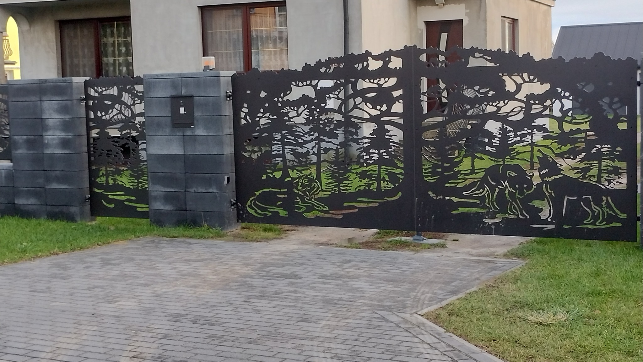 Nowoczesne ogrodzenie z metalu, z wycinanym laserowo wzorem leśnym z motywem wilków, zamontowane na betonowych słupkach. Brukowany podjazd i trawnik.