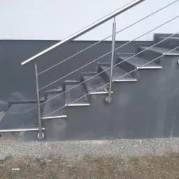 Zewnętrzne schody wyłożone ciemnymi płytkami z metalowym wykończeniem krawędzi i balustradą ze stali nierdzewnej, prowadzące do wejścia budynku z dwukolorową elewacją.