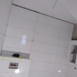 Ściana łazienki w trakcie układania białych, podłużnych płytek; widoczne przymocowane taśmą elementy oraz otwór pod instalację sanitarną.