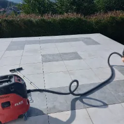 Czyszczenie szarych płytek tarasowych na zewnątrz przy użyciu czerwonego odkurzacza przemysłowego Flex z długim wężem, widok na zielone krzewy i wzg&oacute;rza w tle.