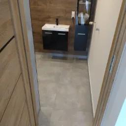 Nowoczesna łazienka z drewnopodobnymi płytkami na ścianie, czarną szafką pod umywalką i wysoką szafką z przeszkloną witryną, widoczna przez otwarte drzwi.