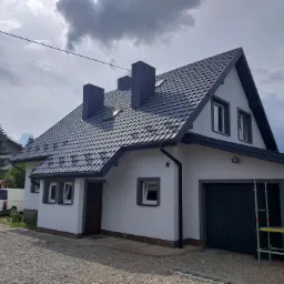 Świeżo pomalowany, biały dom z grafitowym dachem i stolarką okienną, widoczny z perspektywy podwórza z szarym żwirem, drabina oparta o ścianę garażu.