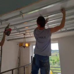 Montaż stelażu sufitu podwieszanego z płyt karton gips, widok na okno i krajobraz za nim, pracownik z wkrętarką i drugi przytrzymujący płytę, w tle wisząca lampa.