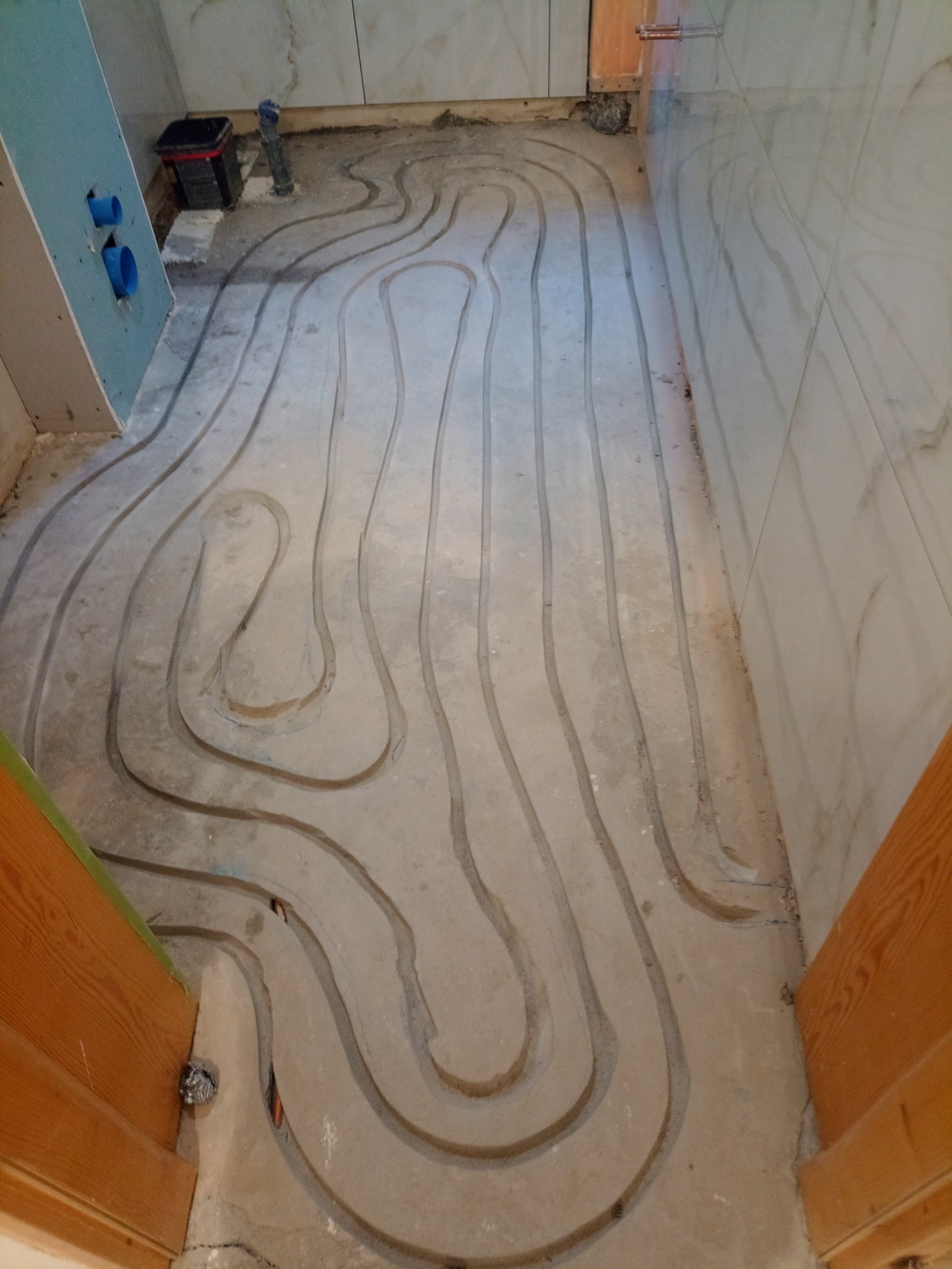 Przygotowanie podłoża pod ogrzewanie podłogowe w łazience, widoczne wyfrezowane rowki w wylewce, fragment ściany z płyt kartonowo-gipsowych oraz częściowo ułożone płytki ścienne.