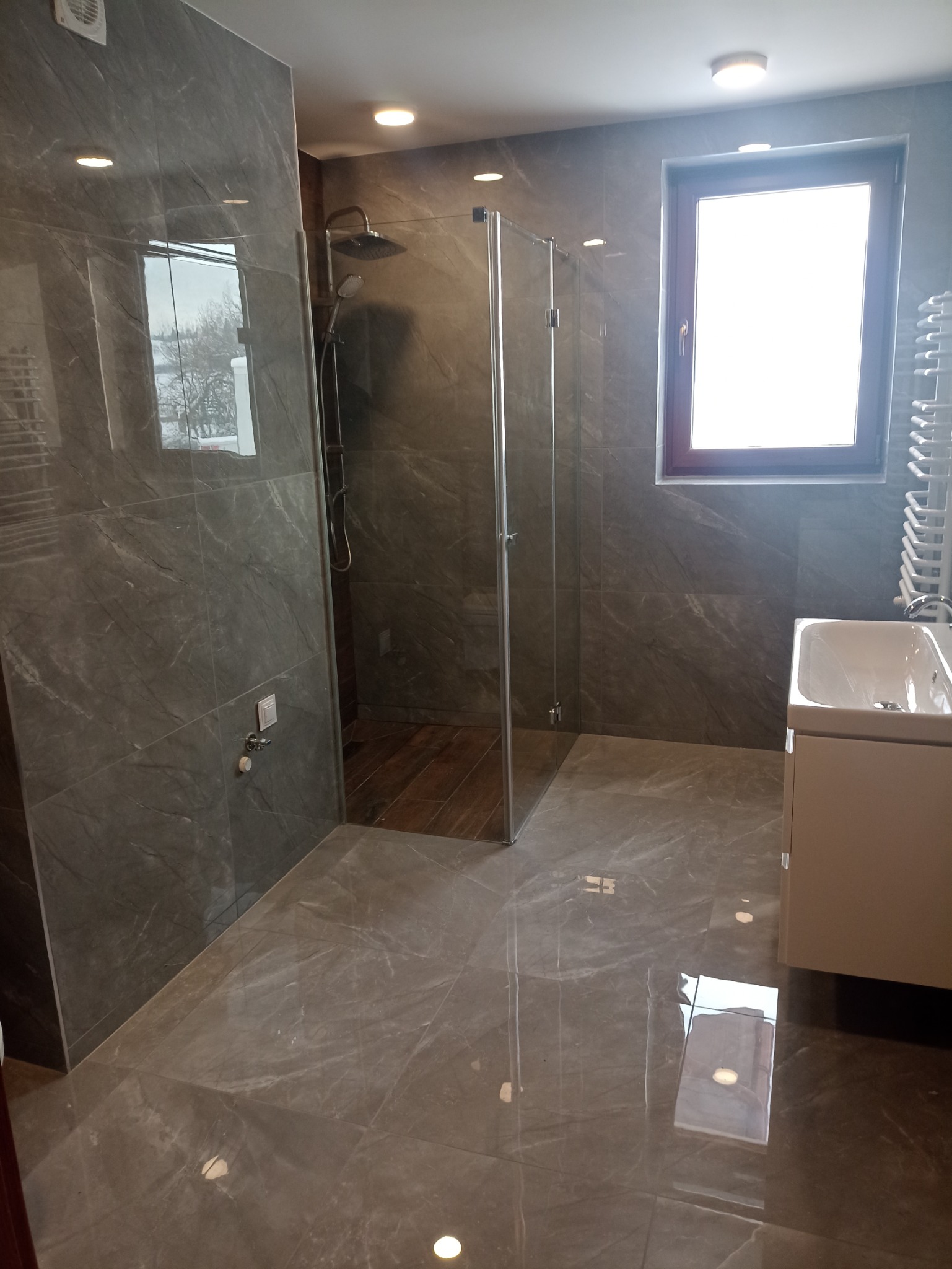 Nowoczesna łazienka z szarymi marmurkowymi płytkami, kabiną prysznicową typu walk-in z deszczownicą, umywalką z szafką i grzejnikiem drabinkowym.
