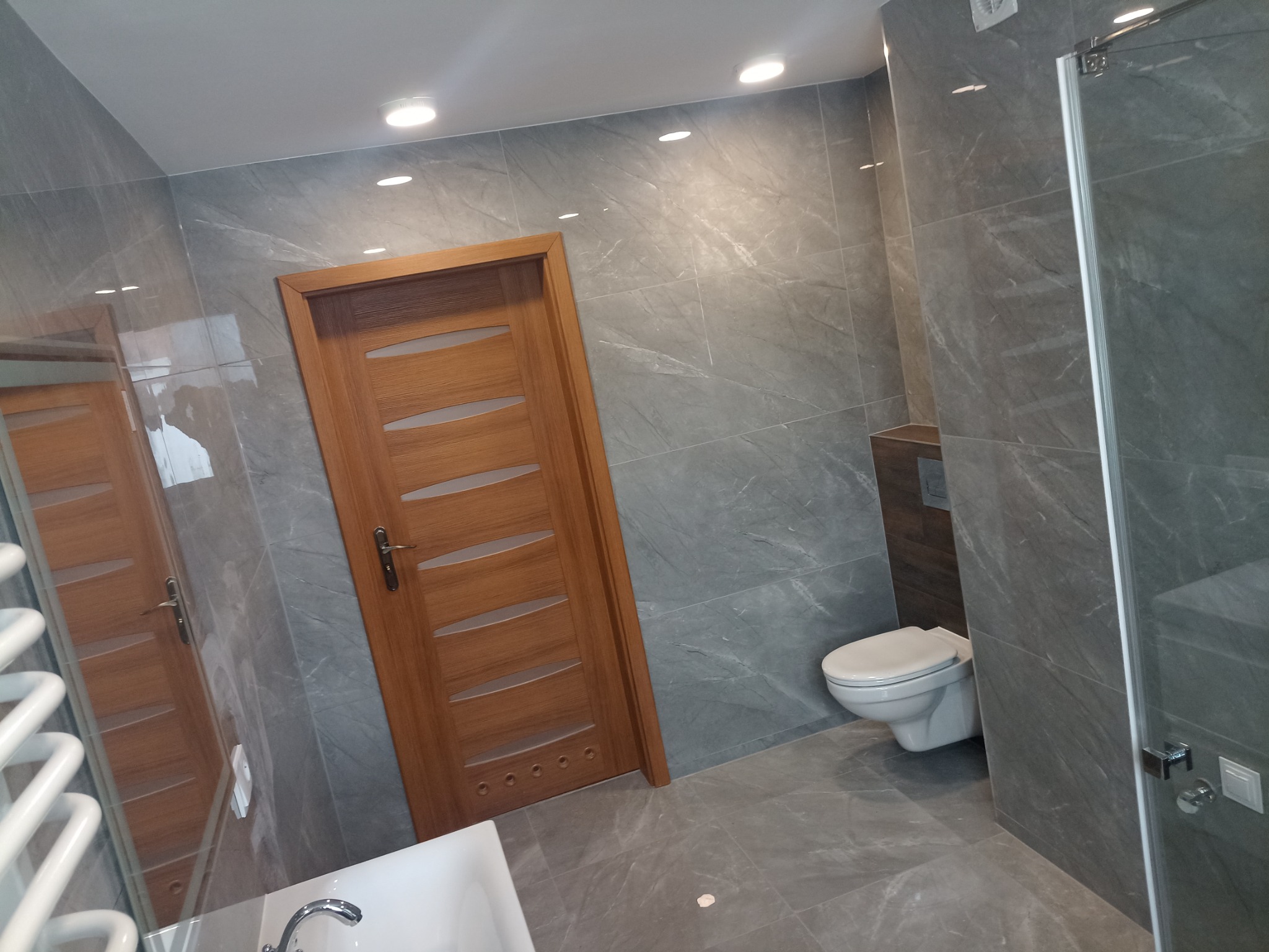 Nowoczesna łazienka z szarymi płytkami imitującymi marmur, drewnianymi drzwiami z poziomymi przeszkleniami, białą toaletą podwieszaną i częściowo widoczną kabiną prysznicową z metalowymi okuciami...