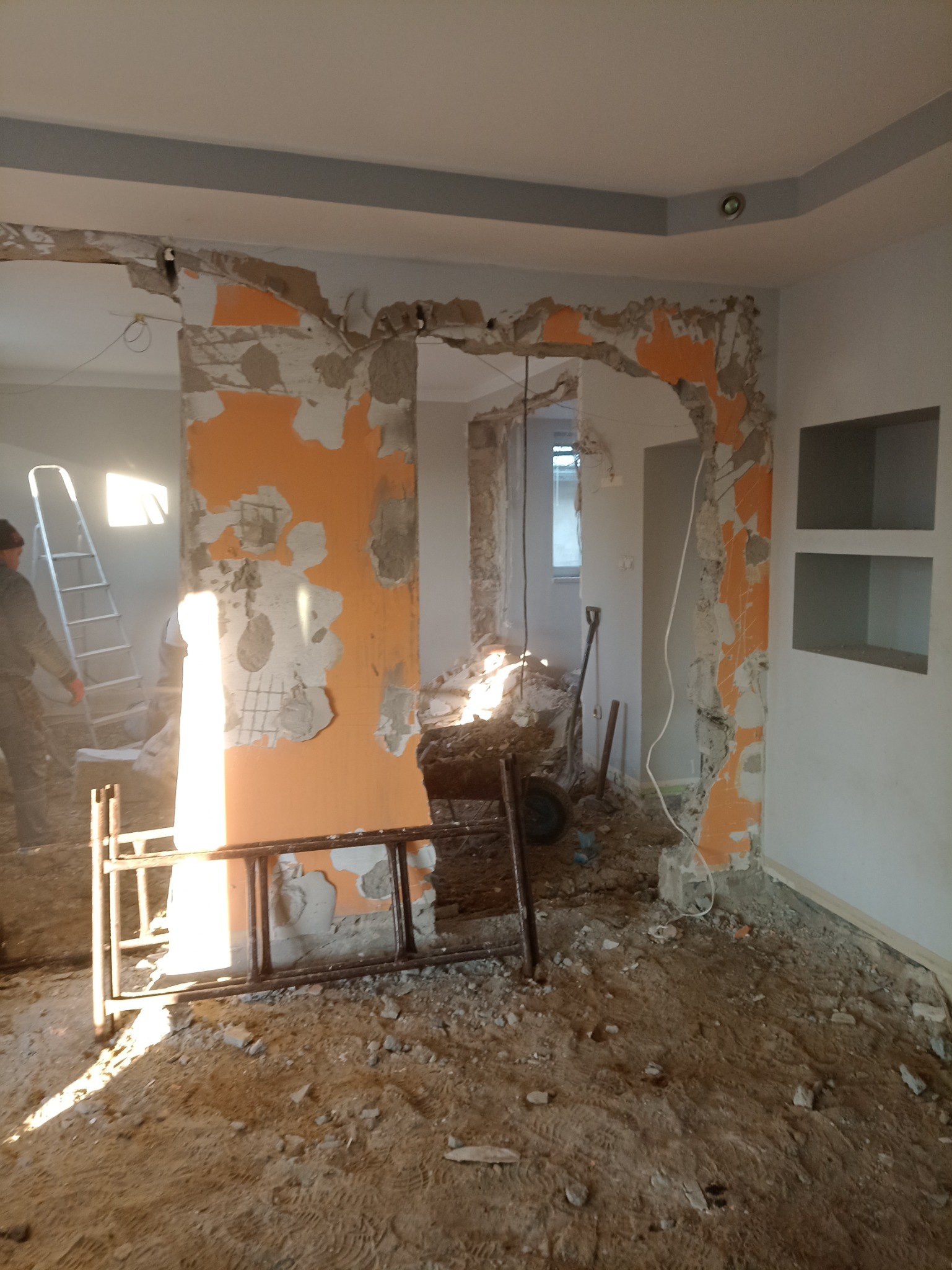 Demontaż ścian działowych w trakcie remontu mieszkania, widoczne fragmenty pomarańczowej farby, gruz na podłodze, drabina i osoba w tle.