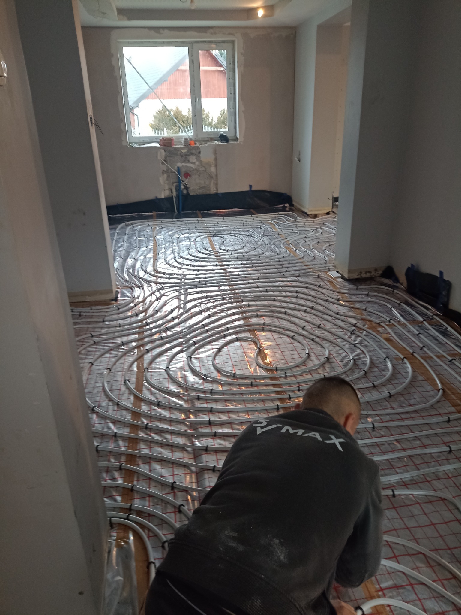 Instalacja ogrzewania podłogowego w pomieszczeniu w trakcie remontu; widoczne rury grzewcze ułożone na macie izolacyjnej, fragment ściany z oknem i pracownik w bluzie z napisem V-MAX.
