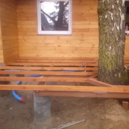 Drewniana konstrukcja tarasu budowana wokół pnia drzewa, widoczne betonowe podpory i fragment elewacji budynku z drewnianą okładziną.