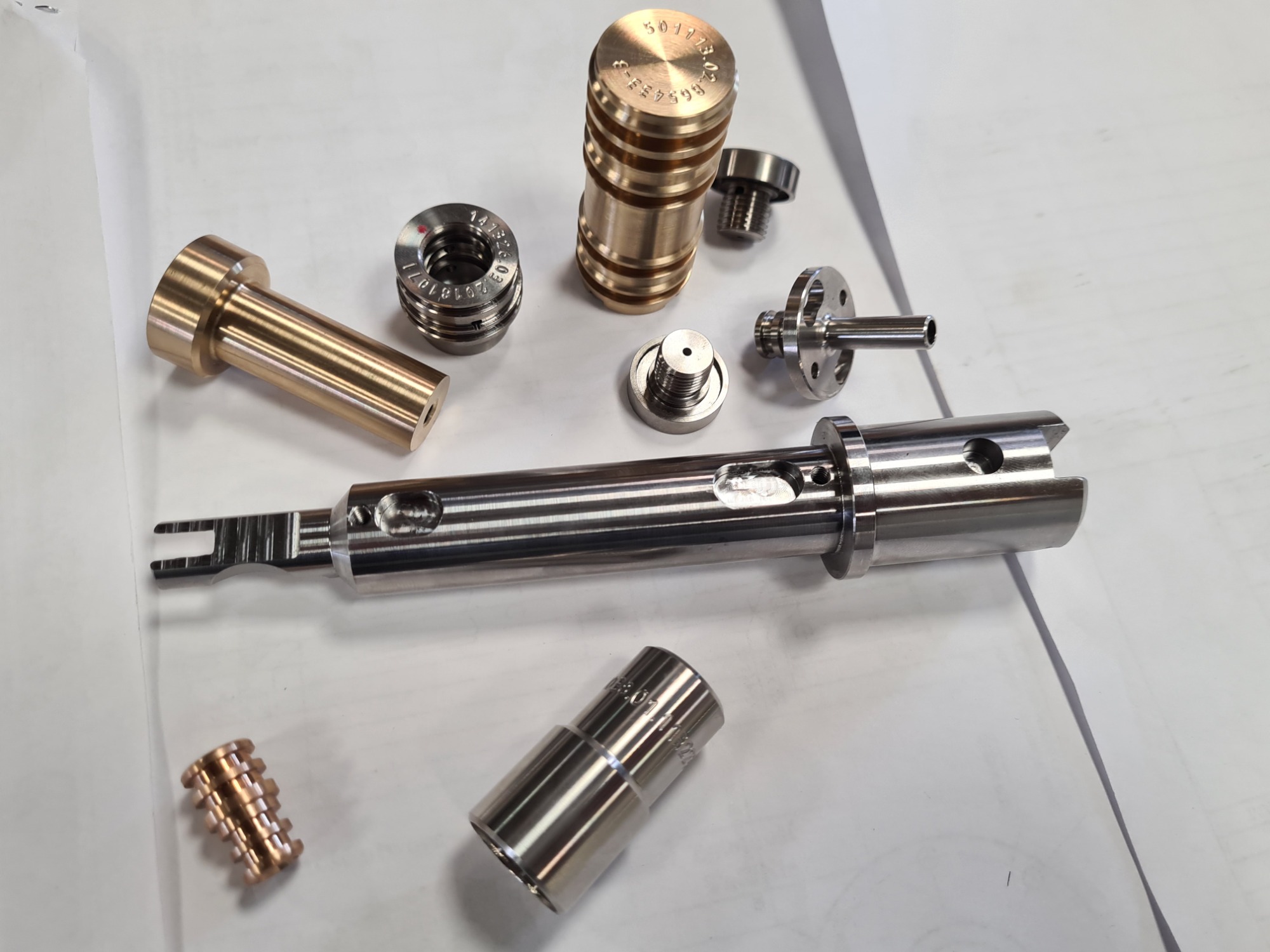 Precyzyjnie wykonane elementy metalowe z różnymi gwintami i otworami, położone na białym tle, widoczne numery seryjne i oznaczenia na powierzchniach.