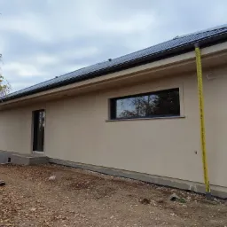 Wykończona elewacja domu jednorodzinnego w odcieniach beżu, z widocznym fragmentem dachu pokrytego ciemną dachówką, żółta rura spustowa i teren wokół budynku pokryty liśćmi.