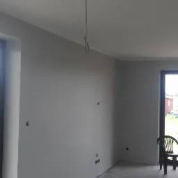 Wnętrze pokoju po malowaniu ścian na szaro, widoczne przygotowane otwory na gniazdka elektryczne, surowe kable zwisające z sufitu i dwa plastikowe krzesła przy oknie balkonowym.