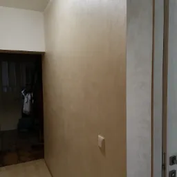 Beżowa ściana z dekoracyjną strukturą, widoczny włącznik światła i fragment drzwi z klamką, w tle ciemne pomieszczenie.