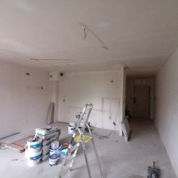 Wykończenie mieszkań - Pokój w trakcie szpachlowania: widoczne worki z gładzią, wiadra z farbą, drabina, ściany pokryte szpachlą, przygotowane do malowania.