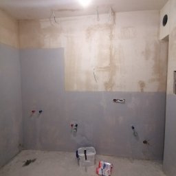 Wykończenie mieszkań - Surowy stan łazienki w trakcie remontu: częściowo położona gładź, widoczne rury wodne, kable elektryczne wystające ze ściany, pudełko i paczka materiałów budowlanych na podłodze.