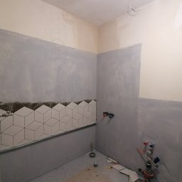 Wykończenie mieszkań - Wnętrze łazienki w trakcie remontu: częściowo ułożone białe, geometryczne płytki na ścianie, surowe ściany przygotowane do dalszych prac wykończeniowych, widoczne narzędzia glazurnicze i instalacja...
