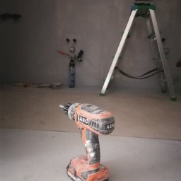 Wykończenie mieszkań - Brudna wiertarko-wkrętarka AEG na podłodze w surowym pomieszczeniu, w tle drabina aluminiowa i puszka z pianką montażową.