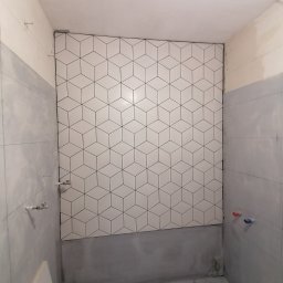 Wykończenie mieszkań - Ściana łazienki wyłożona białymi, sześciennymi płytkami z czarnym obrysem, widoczne przyłącza wodne i ślady prac remontowych na podłodze.