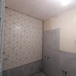Wykończenie mieszkań - Remont łazienki w trakcie: ściana częściowo wyłożona białymi płytkami z czarnym geometrycznym wzorem kostek, surowa ściana przygotowana do dalszych prac, wystające rury instalacji wodnej.