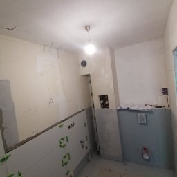 Wykończenie mieszkań - Wnętrze łazienki w trakcie remontu: częściowo ułożone jasnoszare płytki ścienne z krzyżykami poziomującymi, widoczne instalacje, surowe ściany i podłoga, przygotowywana do dalszych prac...