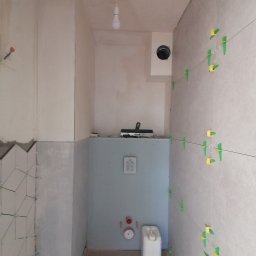 Wykończenie mieszkań - Wnętrze łazienki w trakcie remontu: częściowo ułożone płytki na ścianach, z użyciem poziomicy i klipsów dystansowych, widoczna instalacja wodna i kanister, surowa ściana w tle.