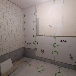 Wykończenie mieszkań - Wnętrze łazienki w trakcie układania płytek, widoczne płytki z geometrycznym wzorem na ścianie, poziomice laserowe, krzyżyki dystansowe i przygotowane otwory na instalacje.