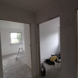 Wykończenie mieszkań - Surowe wnętrze w trakcie przygotowań do malowania: widoczne szpachlowane ściany, drabina aluminiowa pod oknem i wiadro z farbą oraz narzędzia malarskie w drugim pomieszczeniu.