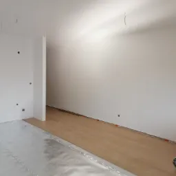 Podkład pod panele z folii aluminiowej oraz częściowo ułożone panele podłogowe w jasnym odcieniu drewna w nowym, pustym pomieszczeniu z białymi ścianami i wystającymi kablami ze ściany.