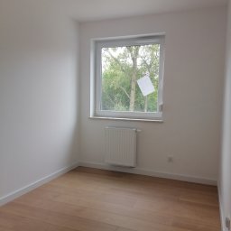 Wykończenie mieszkań - Pusty pokój po remoncie: świeżo pomalowane białe ściany, ułożona jasna podłoga z paneli, biały grzejnik pod oknem z widokiem na drzewa.