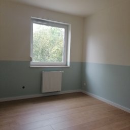 Wykończenie mieszkań - Pomalowane na dwa kolory wnętrze pokoju z drewnianą podłogą, białym grzejnikiem i oknem z widokiem na zieleń. Dolna część ścian w odcieniu pastelowej zieleni, górna kremowa. Z sufitu zwisają kable.