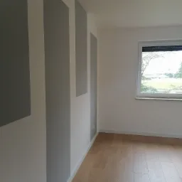 Wnętrze pokoju po malowaniu ścian w pionowe pasy szarości i bieli, z drewnianą podłogą i oknem z widokiem na zieleń.