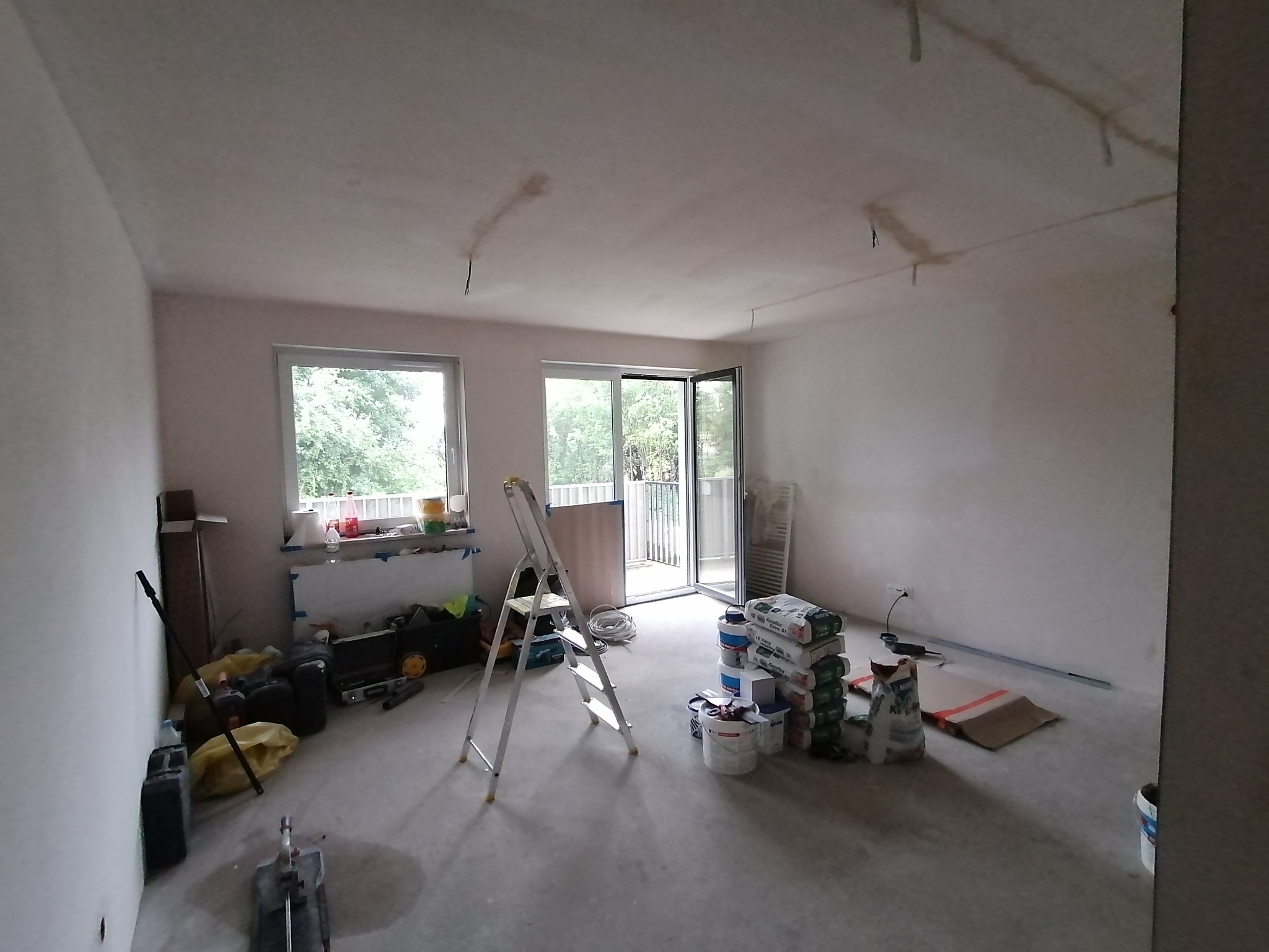 Wnętrze pokoju w trakcie remontu: widoczne worki z zaprawą, drabina aluminiowa, narzędzia malarskie, ślady po instalacjach na suficie przed położeniem gładzi.