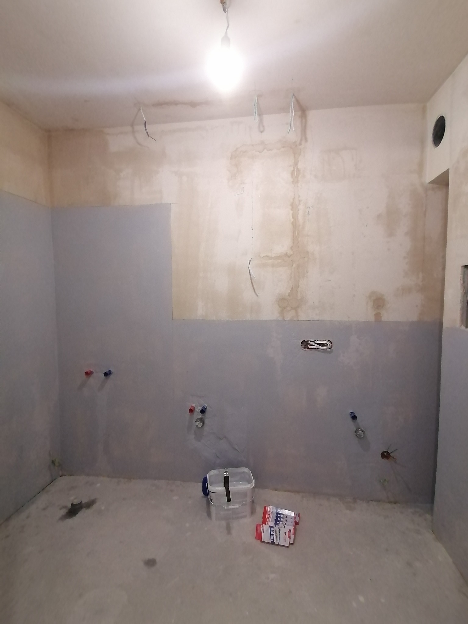 Surowy stan łazienki w trakcie remontu: częściowo położona gładź, widoczne rury wodne, kable elektryczne wystające ze ściany, pudełko i paczka materiałów budowlanych na podłodze.