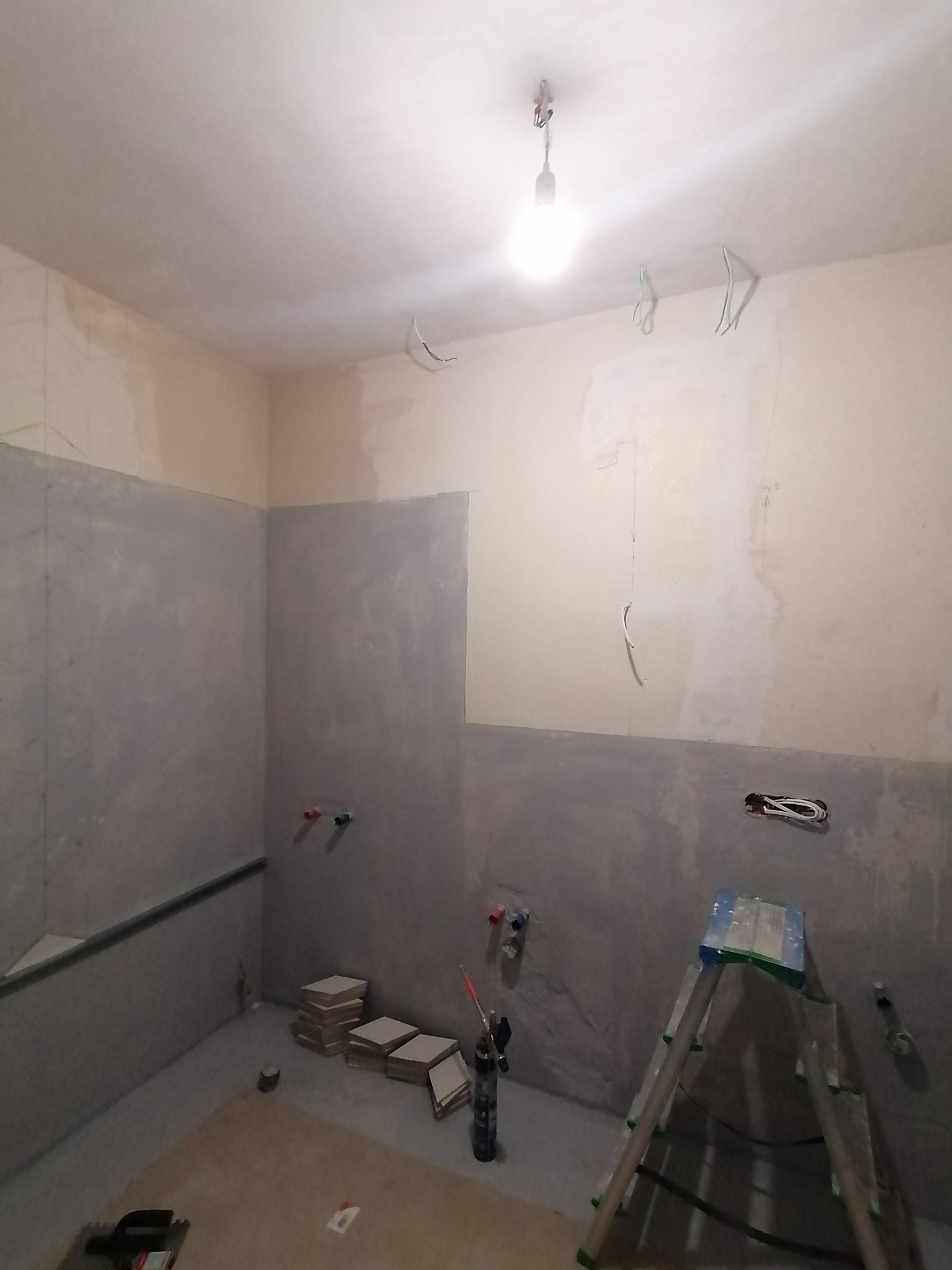 Wnętrze łazienki w trakcie prac remontowych: częściowo położone szare płytki, przygotowane do położenia kolejne, widoczne wystające kable, drabina i narzędzia.
