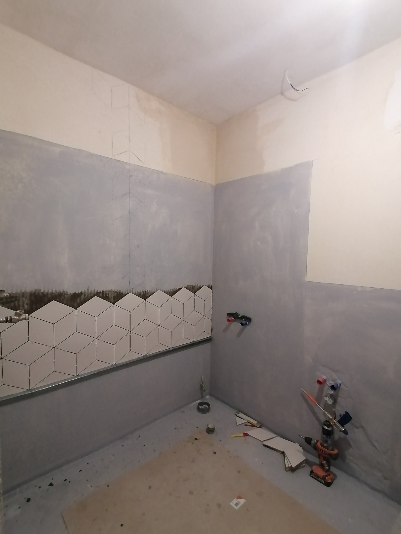 Wnętrze łazienki w trakcie remontu: częściowo ułożone białe, geometryczne płytki na ścianie, surowe ściany przygotowane do dalszych prac wykończeniowych, widoczne narzędzia glazurnicze i instalacja...
