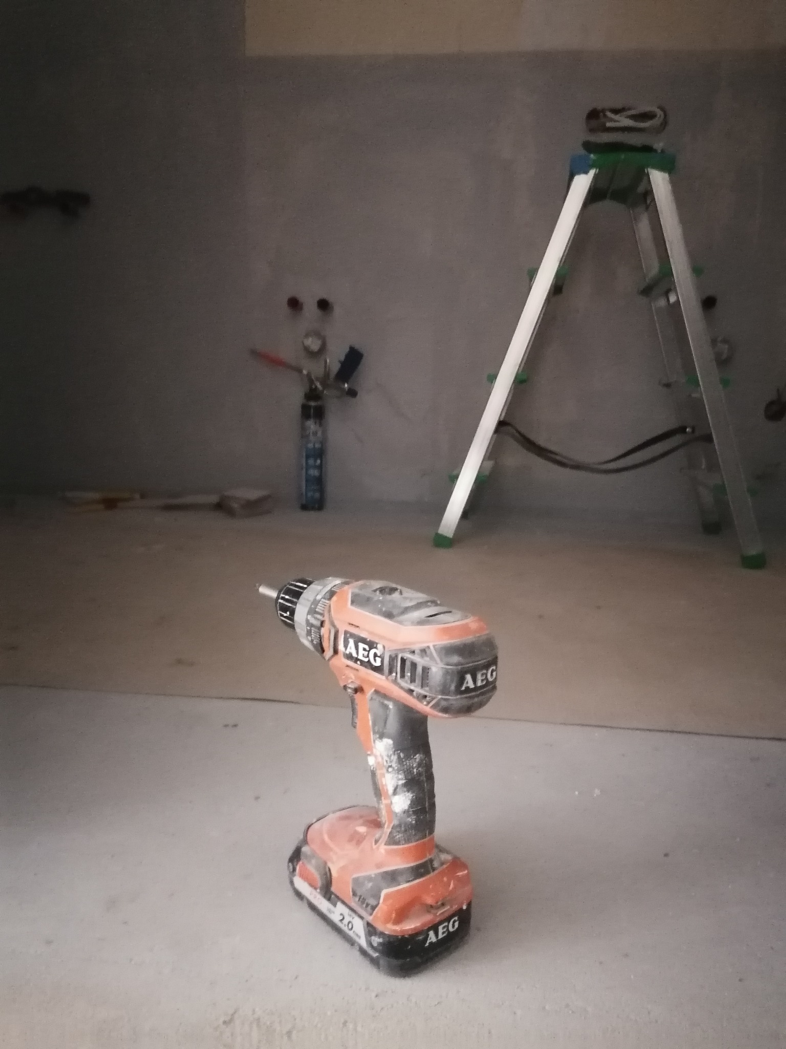 Brudna wiertarko-wkrętarka AEG na podłodze w surowym pomieszczeniu, w tle drabina aluminiowa i puszka z pianką montażową.