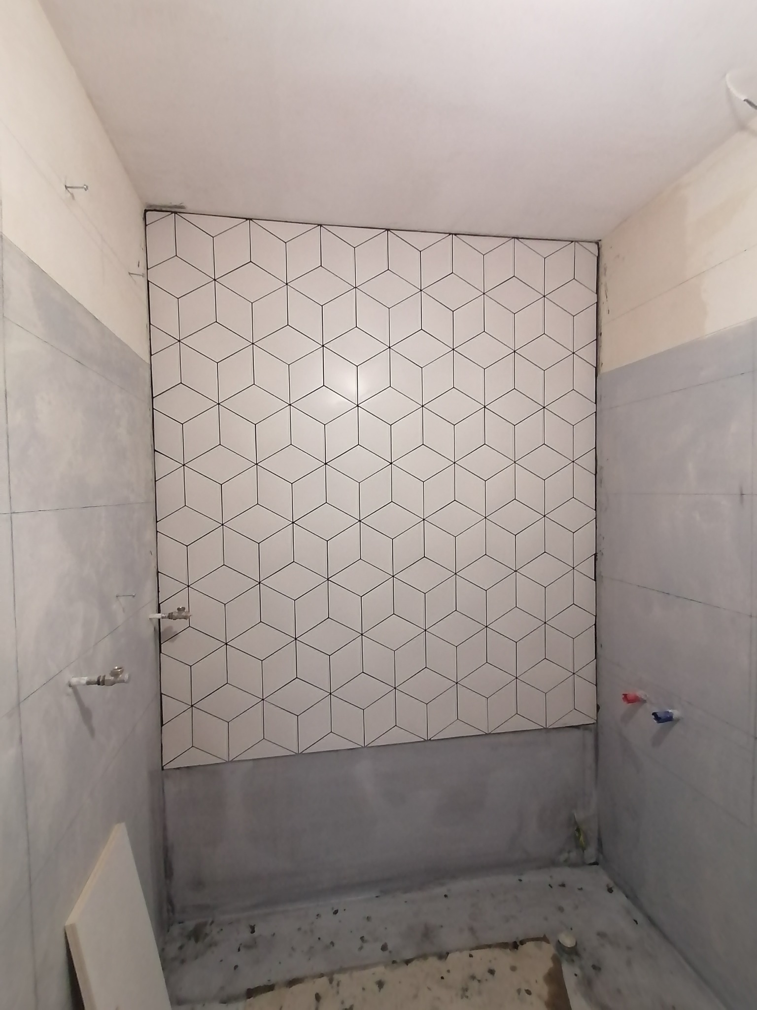 Ściana łazienki wyłożona białymi, sześciennymi płytkami z czarnym obrysem, widoczne przyłącza wodne i ślady prac remontowych na podłodze.