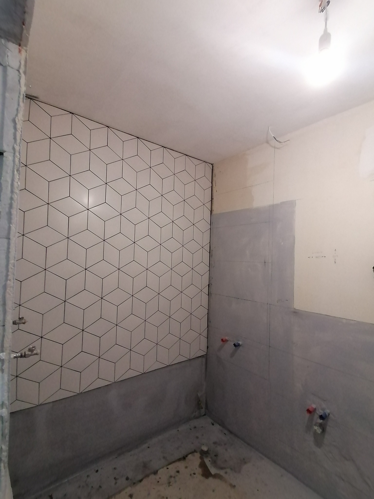 Remont łazienki w trakcie: ściana częściowo wyłożona białymi płytkami z czarnym geometrycznym wzorem kostek, surowa ściana przygotowana do dalszych prac, wystające rury instalacji wodnej.