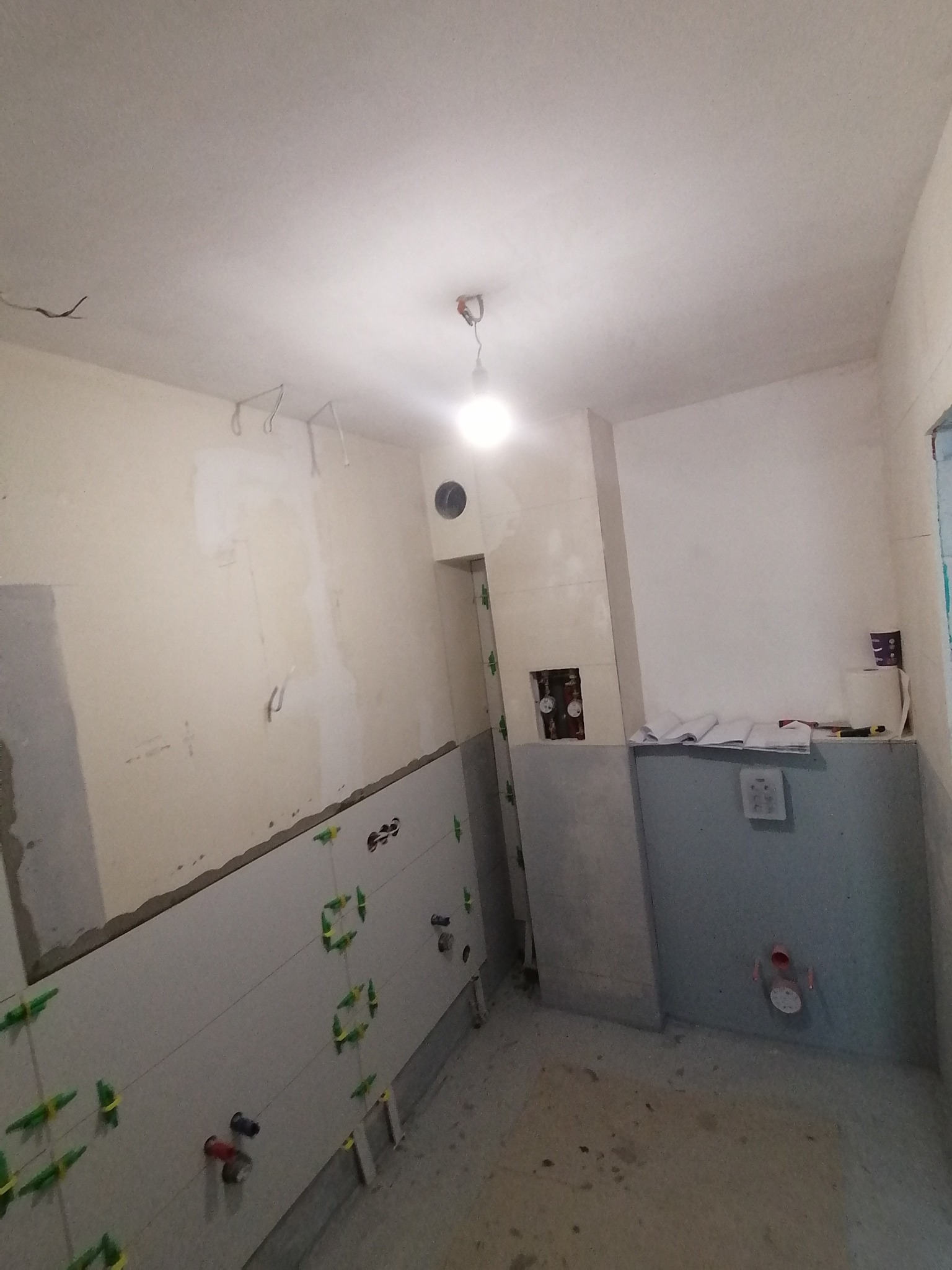 Wnętrze łazienki w trakcie remontu: częściowo ułożone jasnoszare płytki ścienne z krzyżykami poziomującymi, widoczne instalacje, surowe ściany i podłoga, przygotowywana do dalszych prac...